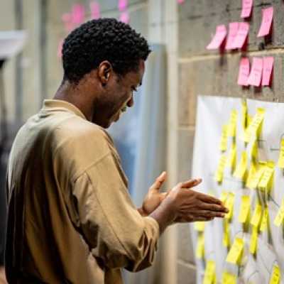 A Black man with a short afro looks at post-it notes on a wall. He is smiling at something he has read. 