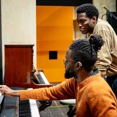 Two people are looking at some sheet music at a piano. One is sitting and holding the music as if they are about to play it, The other is standing, leaning over a chair. 