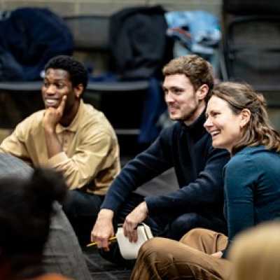 A group of people sit cross-legged on the floor of a rehearsal room smiling, including a Black man of average height with short Afro hair, a white man of average height with light brown hair and a tall woman with brown hair. There are some chairs stacked up against the wall in the background. 