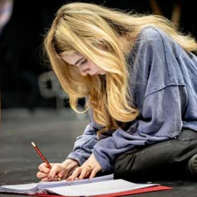 A petite, pale person with chest-length blonde hair sits cross-legged on the floor, marking papers in a folder with a pencil. She wears a blue sweater.