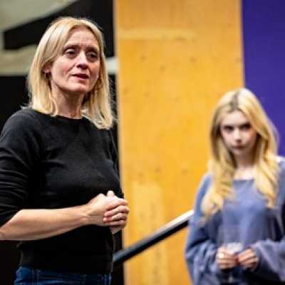 A tall woman with blonde hair and green eyes wears a black jumper and holds her hands clasped in front of her. Out of focus in the background is a second person, who is petite and pale with chest-length blonde hair. 