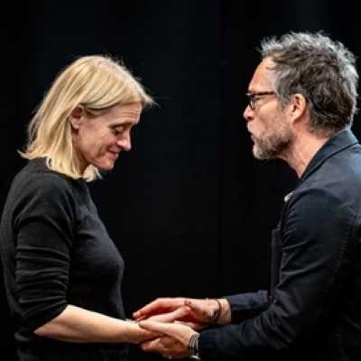 A white man holds the hands of a blonde woman who is standing opposite him. He is talking to her and she is looking down at their hands.