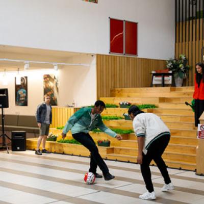 A group of people playing football inside. There are plants covering a wooden staircase, and a tv screen shows a picture of a tall building.