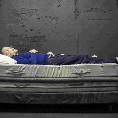 Hans Kesting wearing blue jumper and jeans, lying on a bed. Kesting's clothes are lightly covered in dirt. At the foot of the bed on the floor there is a white metal canister, standing upright in a white stand.