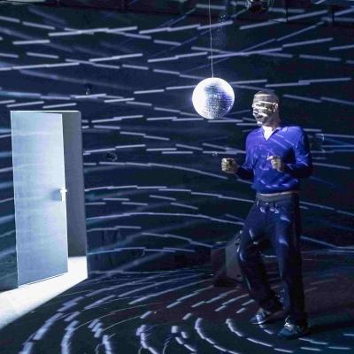 Hans Kesting dancing in front of a disco ball reflecting shards of light across the room. In the background, a TV is visible on the floor, and there is an open doorway.