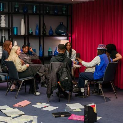 A group of people sitting in chairs in a circle, in the corner of a large room with with grey walls, blue carpet and vases on shelves in the background, and a red curtain. There are pieces of paper, files and bags on the floor. The people smile and laugh as they talk to each other. 