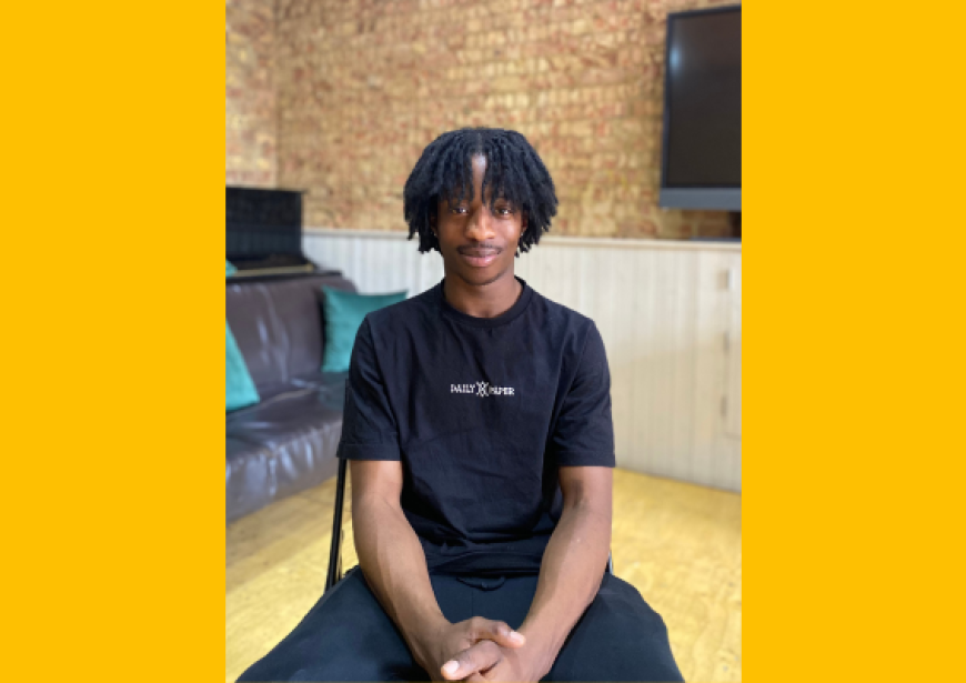 A headshot of a person with an afro wearing a black t-shirt and trousers.