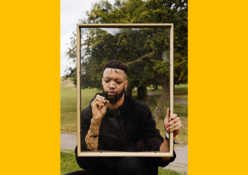 A person with short dark hair and a beard wearing a black top stands holding a frame in front of their face. 