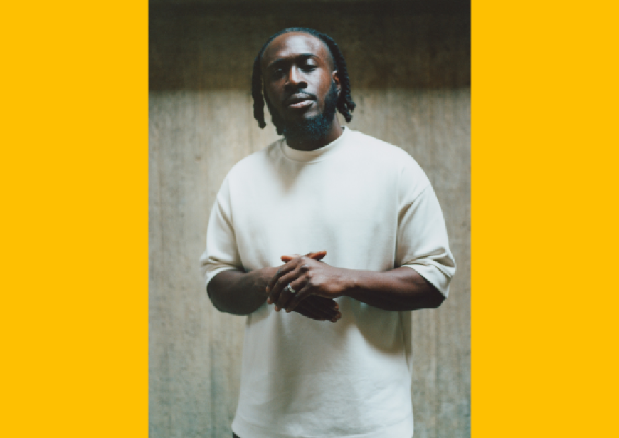 A person with dreads wearing a white t-shirt stands with hands clasped. 