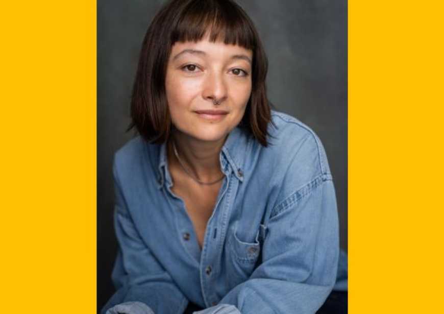 A person with short dark hair with full fringe wearing a blue long sleeved jean shirt and a septum ring.