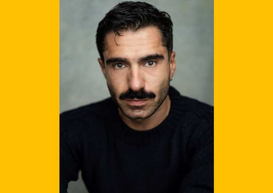 A headshot of a person with short dark hair and a moustache wearing a black long sleeved t-shirt