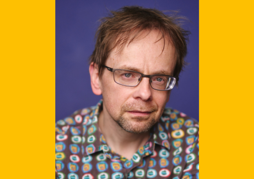 A headshot of a person with short brown hair wearing a checked shirt and glasses