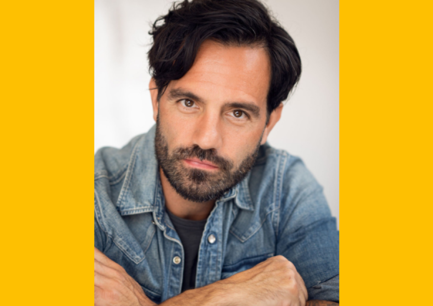 A headshot of a person with short dark hair, a short beard and a moustache wearing a long sleeved denim shirt