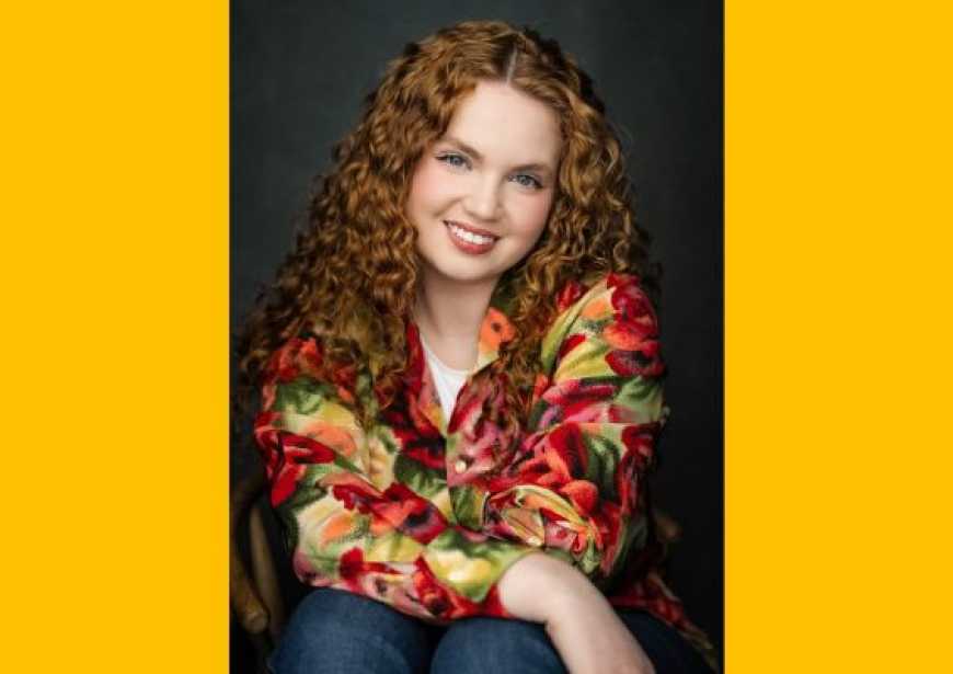 A person with long red curly hair wearing a colourful blouse smiling at the camera