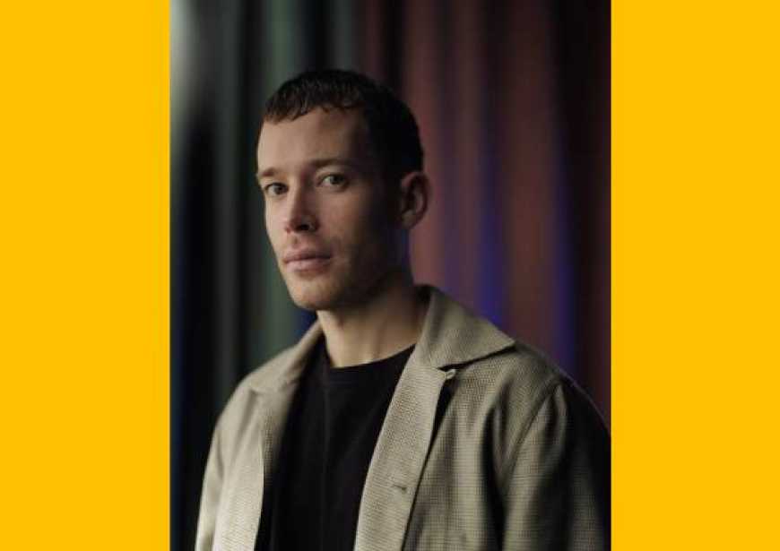 A white man with dark closely cropped hair wearing a beige long sleeved-shirt open over a black t-shirt