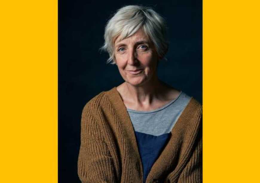 A headshot of a white woman with short gray hair wearing a brown cardigan over a gray and blue t-shirt