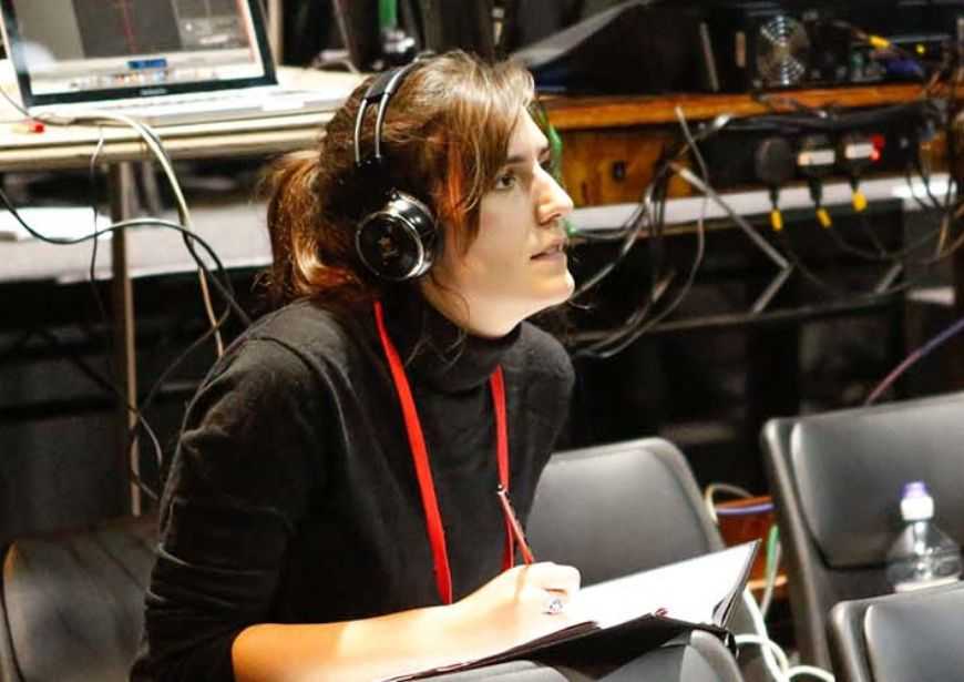 An assistant director with brown hair in a ponytail, wearing a black turtleneck shirt and black headphones, taking notes in front of a sound desk in rehearsals.