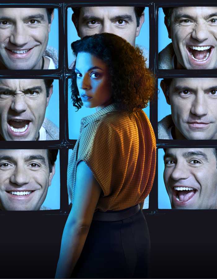 A series of 9 TV screens stacked in columns of 3 show a person's face with different expressions on a blue background. In front of the TV screens stands a person with shoulder length curly hair wearing a yellow blouse turning over their left shoulder to look towards the camera. 
