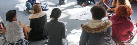 the backs of a group of people sitting on the floor surrounded by paper