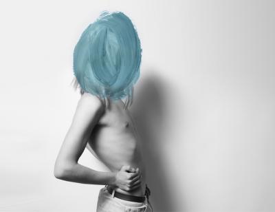 A black and white image of an androgynous person wearing jeans and no top stands side on to the camera. Their face is not visible - the image has thick brush strokes of turquoise paint covering from their shoulders up. Their long blonde hair is just visible beneath the edges of the brush strokes.