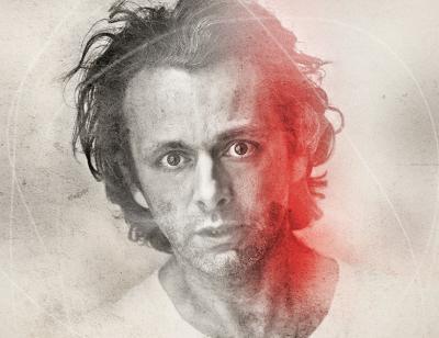 A black and white faded portrait of Michael Sheen, with a red splash on onside of his face 