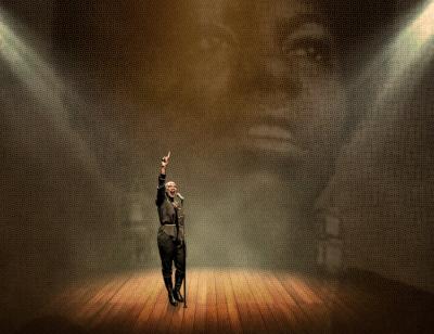 Josette Bushell-Mingo stands on stage with a mic on a stand under a spotlight with her right arm raised in front of a faded portrait on Nina Simone on a backdrop