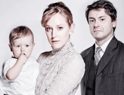 A woman and man stand together holding a baby, staring into the camera. They are all wearing period dress and look serious. The colours in the photo are muted.