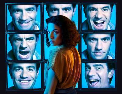 A series of 9 TV screens stacked in columns of 3 show a person's face with different expressions on a blue background. In front of the TV screens stands a person with shoulder length curly hair wearing a yellow blouse turning over their left shoulder to look towards the camera. 